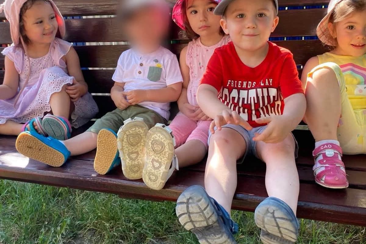 Copii așezați pe o bancă de lemn, la umbră, în curte